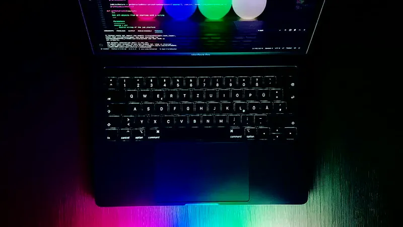 A MacBook laptop open in the dark with four glowing RGB LED juggling balls resting beside it, the balls' colors reflecting off the desk